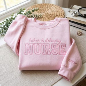 May include: A light pink sweatshirt with the words "labor & delivery NURSE" embroidered on the front. The word "NURSE" is in a gingham pattern. The sleeve cuff has the name "Sarah" embroidered on it.