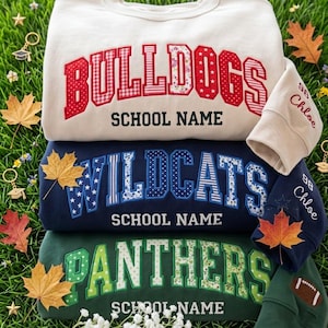 May include: A stack of three crewneck sweatshirts in cream, navy blue, and dark green. Each features large, multi-patterned fabric applique letters spelling BULLDOGS, WILDCATS, or PANTHERS, with SCHOOL NAME printed below. Sleeves show embroidered text.
