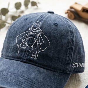 May include: A navy blue baseball cap with a white embroidered design of a person carrying a child on their shoulders. The name "ETHAN" is embroidered on the side. The cap has a curved brim and a button on top.