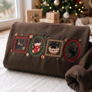 May include: A dark brown sweatshirt featuring embroidered designs of four black cats in festive frames. The embroidery includes a cat with a scarf, a cat in a Christmas stocking, a cat in a bowl, and a cat playing with greenery, accented by stars.