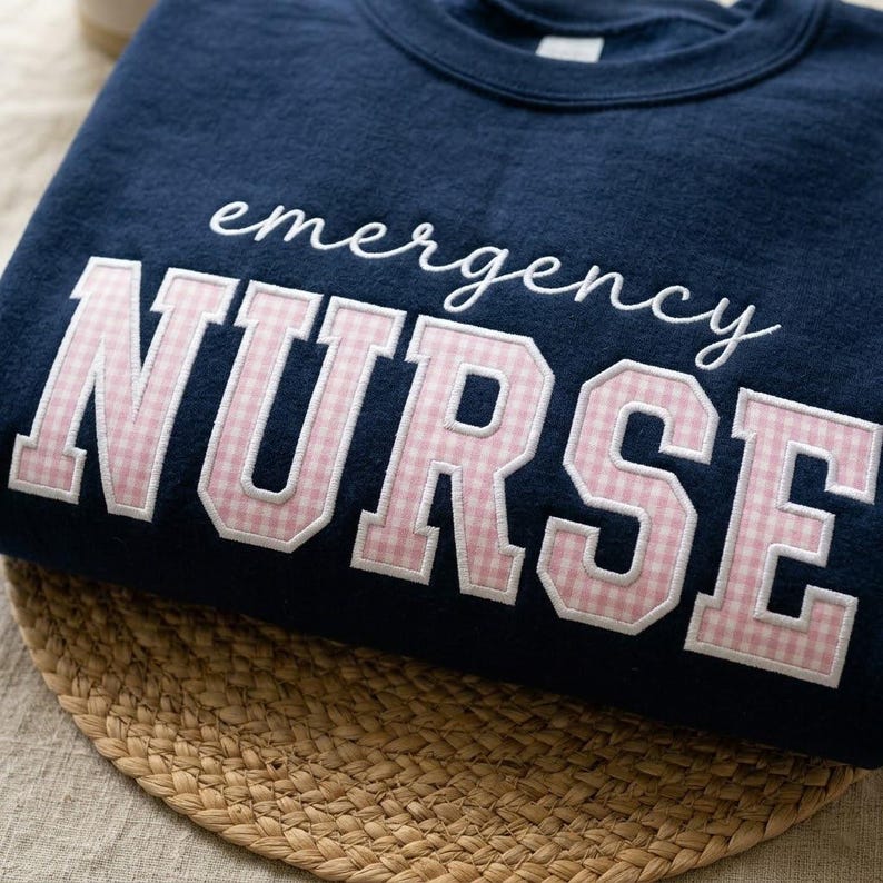 May include: Navy blue sweatshirt with the words "emergency NURSE" in white script and pink gingham letters. The sweatshirt is displayed on a woven mat.