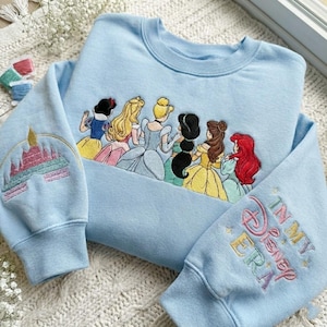 May include: Light blue sweatshirt with embroidered Disney princesses across the chest. The left sleeve features a castle design, and the right sleeve says "In My Disney Era". The sweatshirt is a crew neck style.
