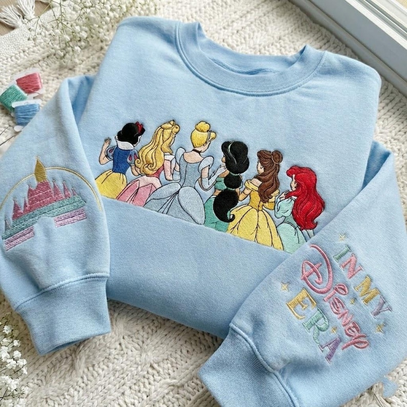 May include: Light blue sweatshirt with embroidered Disney princesses across the chest. The left sleeve features a castle design, and the right sleeve says "In My Disney Era". The sweatshirt is a crew neck style.