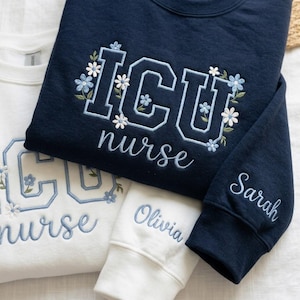 May include: Navy blue and white sweatshirts embroidered with "ICU nurse" and floral accents. One sleeve is embroidered with the name "Sarah", and another with "Olivia". The sweatshirts are a comfortable, casual style.