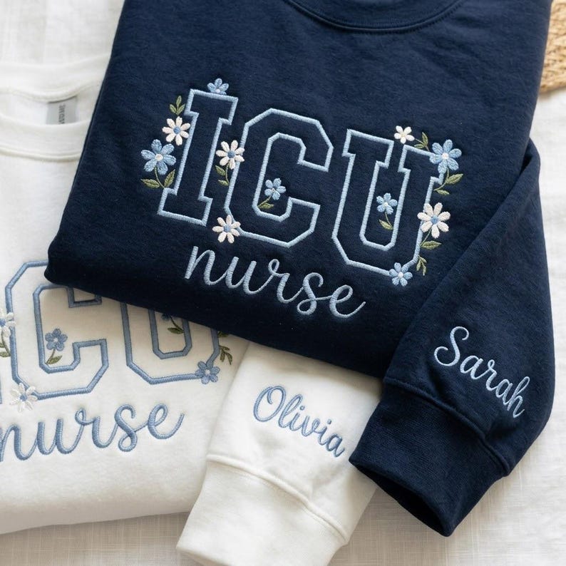 May include: Navy blue and white sweatshirts embroidered with "ICU nurse" and floral accents. One sleeve is embroidered with the name "Sarah", and another with "Olivia". The sweatshirts are a comfortable, casual style.