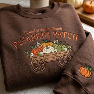 May include: A dark brown crewneck sweatshirt featuring embroidered text that reads Local & Farm Fresh PUMPKIN PATCH above a wooden wagon filled with orange, white, and green pumpkins. A single orange pumpkin is embroidered on the sleeve cuff.
