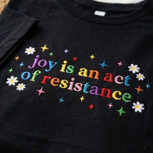 May include: Black t-shirt with embroidered text "joy is an act of resistance" in rainbow colors. The design includes embroidered stars and daisies, creating a colorful and cheerful aesthetic. A casual, comfortable garment.