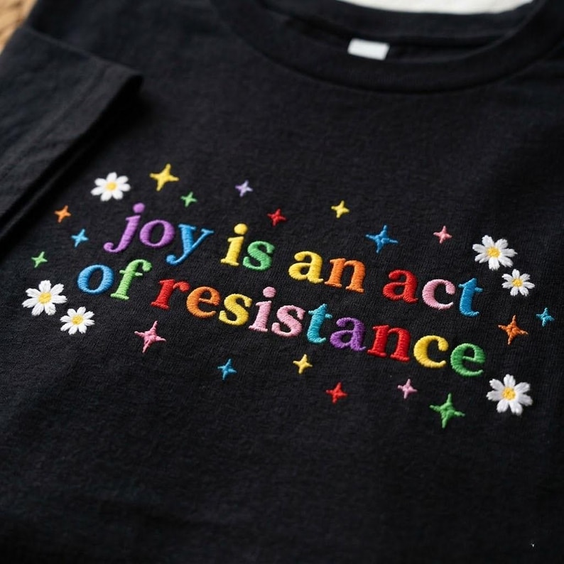 May include: Black t-shirt with embroidered text "joy is an act of resistance" in rainbow colors. The design includes embroidered stars and daisies, creating a colorful and cheerful aesthetic. A casual, comfortable garment.