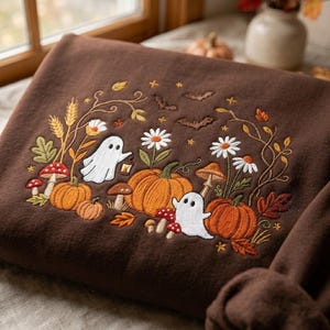 May include: A dark brown sweatshirt featuring detailed embroidery of two white ghosts, orange pumpkins, red and brown mushrooms, white daisies, wheat stalks, and small brown bats arranged in a decorative autumn-themed scene across the front.