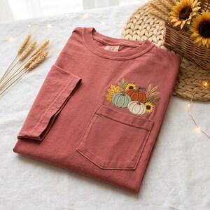 May include: A terracotta-colored short-sleeve t-shirt featuring a chest pocket. The pocket is decorated with an embroidered design of two pumpkins, one light green and one white, surrounded by yellow sunflowers, wheat stalks, and autumn leaves.