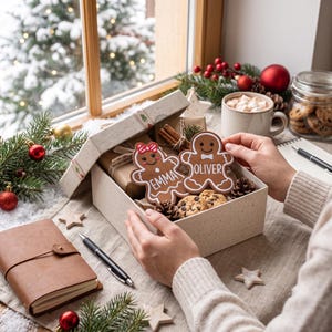 Puede incluir: Una caja de regalo de color claro que contiene dos galletas con forma de hombre de jengibre con detalles de glaseado blanco y los nombres Emma y Oliver escritos. La caja también incluye pequeñas galletas con chispas de chocolate, piñas y canela.