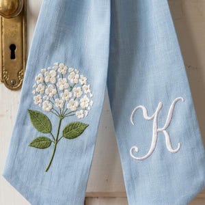 Op de afbeelding: Lichtblauw linnen handdoek met geborduurde witte hortensiabloemen en groene bladeren. De handdoek heeft ook een geborduurde witte letter "K". De handdoek hangt aan een witte deur.