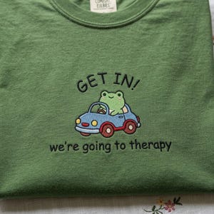May include: Green crew neck sweatshirt with embroidered design. The design features a green frog driving a blue car with the text "GET IN!" above and "we're going to therapy" below.