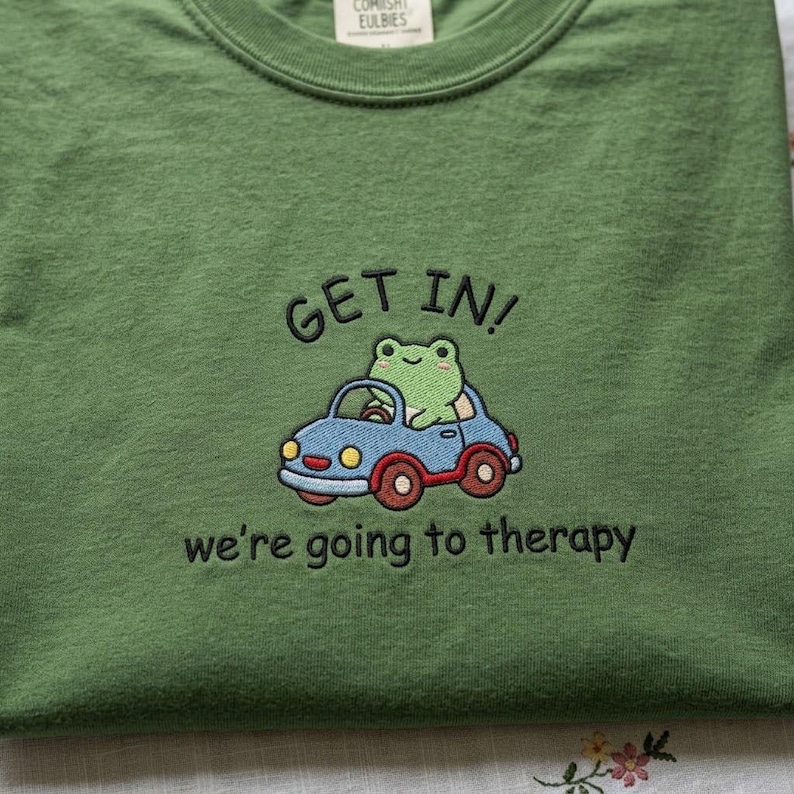 May include: Green crew neck sweatshirt with embroidered design. The design features a green frog driving a blue car with the text "GET IN!" above and "we're going to therapy" below.