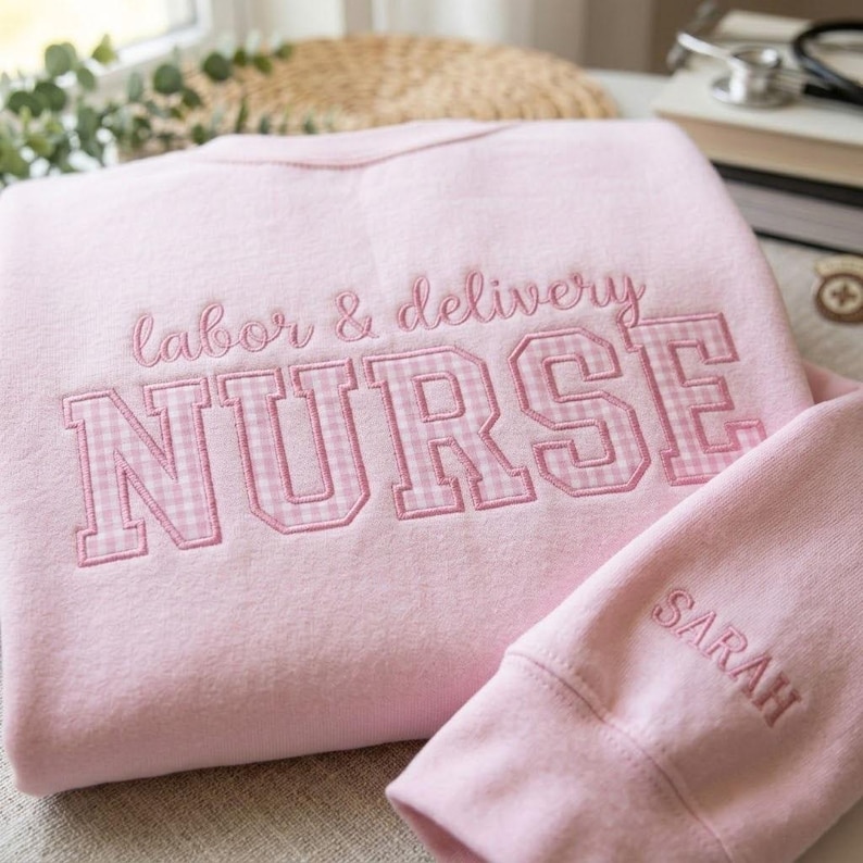 May include: A light pink sweatshirt with the words "labor & delivery NURSE" embroidered in pink. The word "NURSE" is in a gingham pattern. The sleeve has the name "SARAH" embroidered in pink.