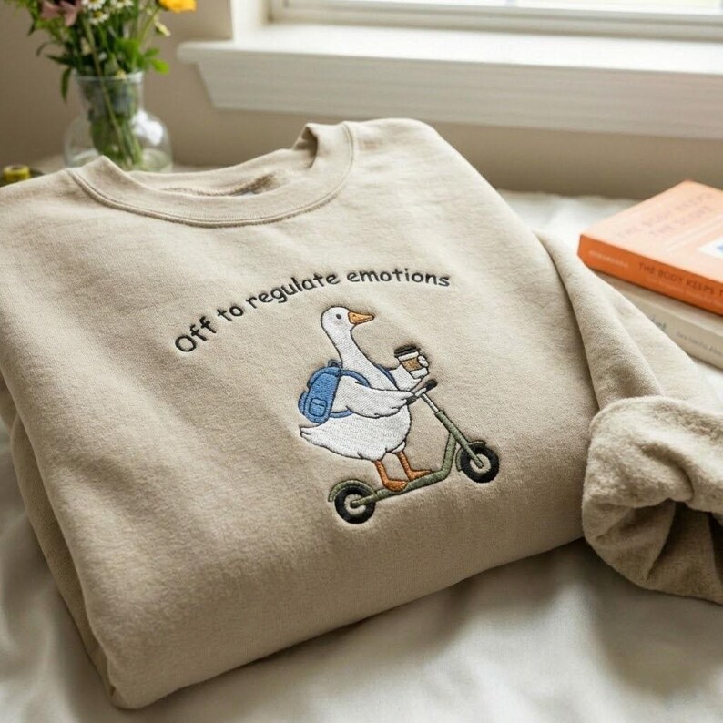 Puede incluir: Sudadera beige con un dise&ntilde;o bordado de un ganso en un scooter, sosteniendo una taza de caf&eacute; y con una mochila. El texto "Off to regulate emotions" est&aacute; encima del ganso. La sudadera est&aacute; doblada sobre una superficie blanca.