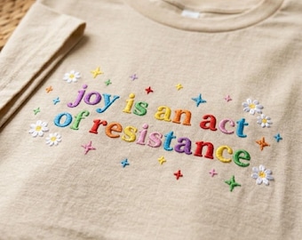 Pode incluir: Camiseta bege de gola redonda com a frase bordada "joy is an act of resistance" em cores do arco-íris. O design inclui flores e estrelas bordadas.