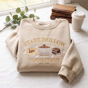 May include: A beige crewneck sweatshirt featuring gold embroidered text that reads Stars Hollow Connecticut Est. 1779. The design includes a central gazebo, a yellow sign with the text Luke's, a stack of books, and small white daisy flowers.
