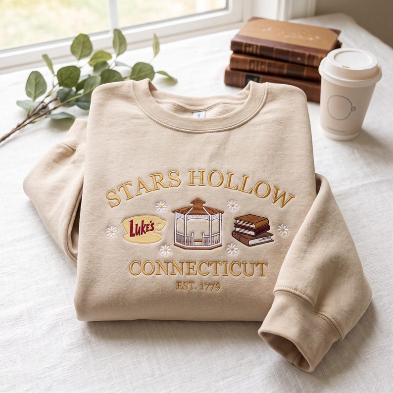 May include: A beige crewneck sweatshirt featuring gold embroidered text that reads Stars Hollow Connecticut Est. 1779. The design includes a central gazebo, a yellow sign with the text Luke's, a stack of books, and small white daisy flowers.