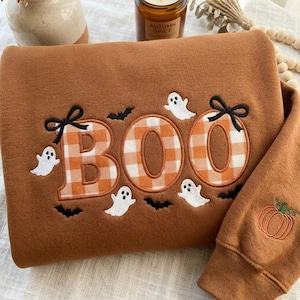 Op de afbeelding: Een bruine sweater met de geborduurde tekst BOO in een oranje en wit ruitpatroon. Het ontwerp bevat zwarte strikjes op de letters B en O, witte spookfiguren en kleine zwarte vleermuizen. Een kleine oranje pompoen staat op de mouw.