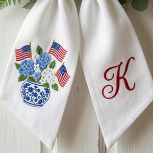 May include: White linen kitchen towels. One towel features an embroidered blue and white vase with blue and white flowers and American flags. The other towel has a red embroidered letter "K".