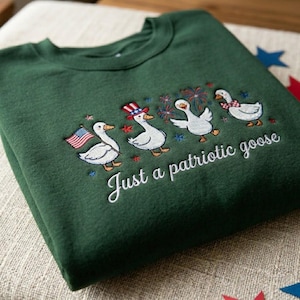 May include: A dark green sweatshirt with an embroidered design of four cartoon geese celebrating with fireworks, an American flag, and a patriotic hat. The text "Just a patriotic goose" is below the geese.