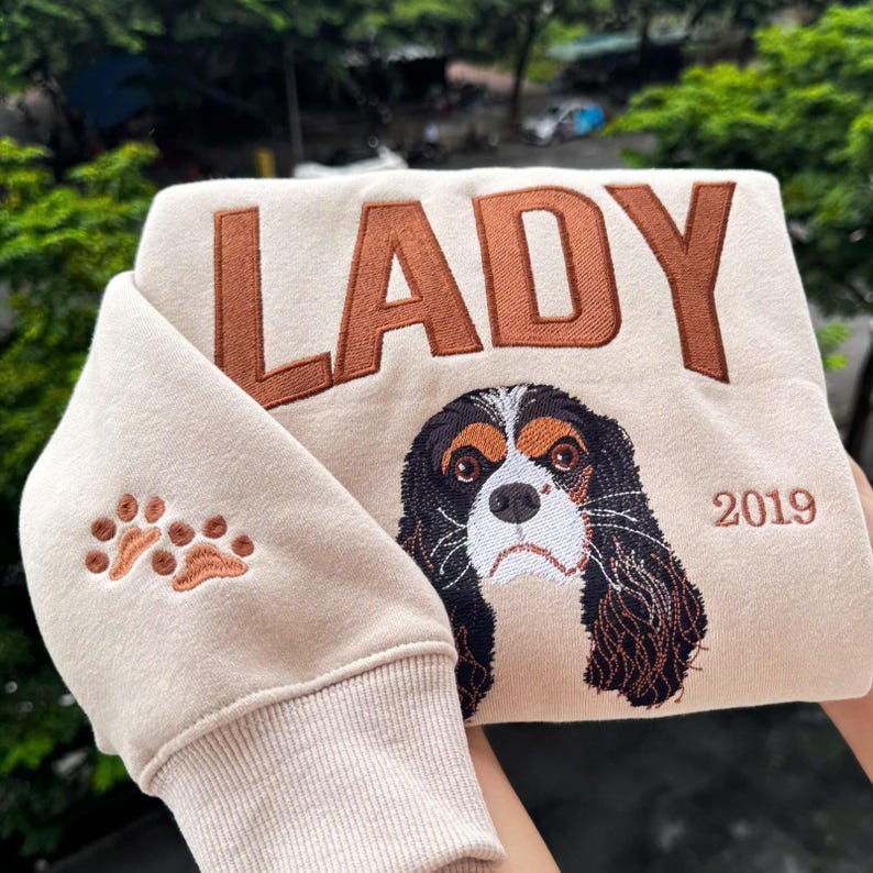 May include: A beige sweatshirt with brown embroidered text reading "LADY". It features a Cavalier King Charles Spaniel design and the year "2019". The left sleeve has paw print embroidery.