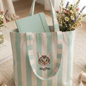 May include: A striped tote bag with a cat face embroidery and the word "Muffins" below it. The bag is filled with a notebook and a bouquet of flowers. The bag has vertical stripes in light green and white.