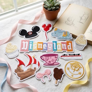 May include: A collection of embroidered patches featuring a Disneyland sign, Mickey ears, a red balloon, Dumbo, a pirate ship, a teacup, a monorail, a cupcake, a pink bow, a chocolate ice cream bar, and a gold circular icon with a smiling face.