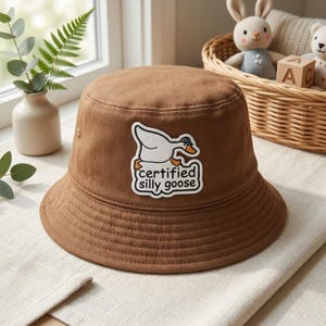 Op de afbeelding: Een bruine bucket hat met een witte geborduurde patch aan de voorzijde. De patch toont een witte gans met een klein blauw hoedje, met de zwarte tekst "certified silly goose" eronder geschreven in een afgerond lettertype.