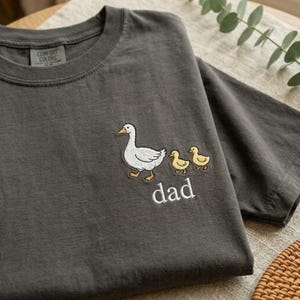 Puede incluir: Camiseta gris oscuro con un dise&ntilde;o bordado de un ganso blanco, dos patitos amarillos y la palabra "dad" en blanco. La camiseta tiene cuello redondo y est&aacute; hecha de un tejido suave.