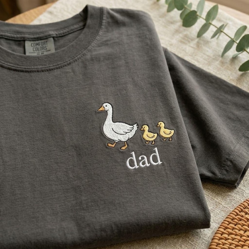 Puede incluir: Camiseta gris oscuro con un dise&ntilde;o bordado de un ganso blanco, dos patitos amarillos y la palabra "dad" en blanco. La camiseta tiene cuello redondo y est&aacute; hecha de un tejido suave.