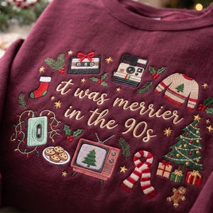 May include: A maroon sweatshirt featuring embroidered holiday-themed 90s icons including a cassette tape, instant camera, television with a tree, and cookies. The text "it was merrier in the 90s" is stitched in gold thread across the center.