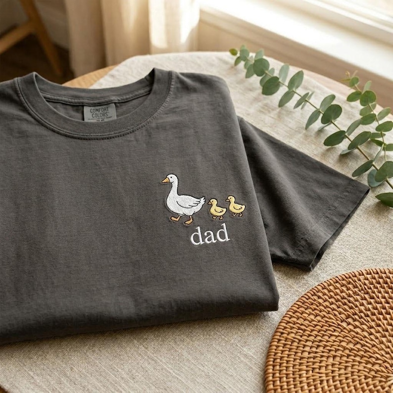 Puede incluir: Camiseta gris oscuro con un bordado de un ganso blanco y dos patitos amarillos, con la palabra "dad" debajo. La camiseta est&aacute; doblada sobre una superficie clara, con una estera de mimbre visible.
