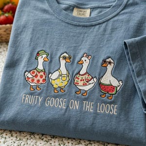 Pode incluir: Camiseta azul Comfort Colors com um design bordado de quatro gansos de desenho animado. Cada ganso está vestido com roupas com tema de frutas, com o texto "FRUITY GOOSE ON THE LOOSE" bordado abaixo. Os gansos usam óculos de sol, chapéus e laços.