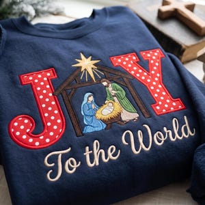 May include: A navy blue sweatshirt featuring an embroidered nativity scene centered between the letters J and Y. The letters are red with white polka dots. A gold star shines above the manger. The text To the World is embroidered below.