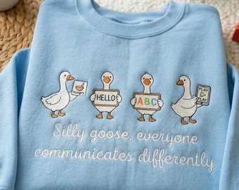 Könnte beinhalten: Hellblaues Sweatshirt mit gestickten Gänsen, die Schilder mit den Wörtern "hungry", "hello", "ABC" und "play" halten. Der Text "Silly goose, everyone communicates differently" ist darunter gestickt.