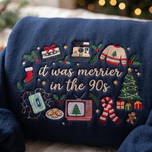 May include: A navy blue sweatshirt featuring embroidered 90s-themed Christmas icons including a cassette tape, instant camera, sweater, stocking, tree, television, cookies, and scarf, with the text "it was merrier in the 90s" in the center.
