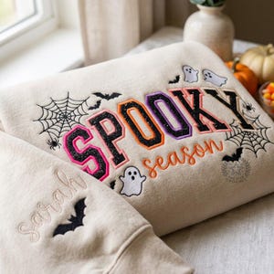 May include: A beige sweatshirt featuring embroidered Halloween designs including black spiderwebs, bats, and white ghosts. The text SPOOKY is in varsity-style letters with black, orange, and purple glitter, while season is in orange script. Sarah is embroidered on the sleeve.