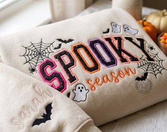Op de afbeelding: Een beige sweatshirt met geborduurde Halloween-ontwerpen, waaronder zwarte spinnenwebben, vleermuizen en witte spoken. De tekst SPOOKY staat in glitterende varsity-letters en season in oranje cursief schrift. De naam Sarah is op de mouw geborduurd.