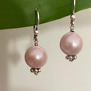 Peut inclure: Boucles d'oreilles pendantes avec de grosses perles rose pâle. Les boucles d'oreilles ont des crochets argentés et un groupe de petites perles argentées à la base. Les boucles d'oreilles sont présentées sur une feuille verte.