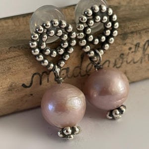 Peut inclure: Une paire de boucles d'oreilles pendantes avec de grosses perles nacrées rose clair. Les boucles d'oreilles ont des cerceaux perlés argentés et de petits accents perlés à la base des perles. Les tiges des boucles d'oreilles sont transparentes.
