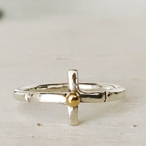 May include: A silver ring with a gold cross design. The cross has a small gold ball in the center.