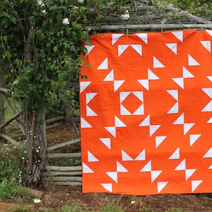 May include: A large orange and white quilt with a geometric pattern of triangles. The quilt is hanging on a wooden fence in a garden setting.