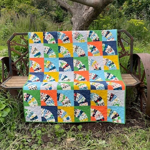 May include: A colorful quilt with a fan-shaped pattern in shades of blue, green, orange, and yellow. The quilt is displayed on a wooden bench with metal wheels.