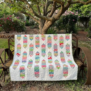 Op de afbeelding: Een wit quilt met een patchwork-ontwerp met kleurrijke, geometrische vormen. Het quilt wordt getoond op een houten bank met een rustiek metalen frame.