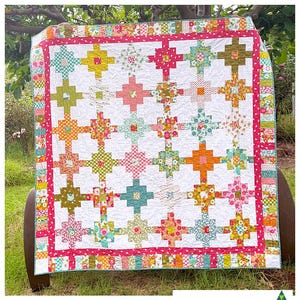 May include: A white quilt with a patchwork design featuring a variety of floral prints in pink, green, orange, and blue. The quilt is bordered with a pink and white striped fabric and a teal and white striped fabric. The quilt is displayed on a wooden chair in a garden setting. The text "KATE HENDERSON QUILTS" is visible at the bottom of the image.