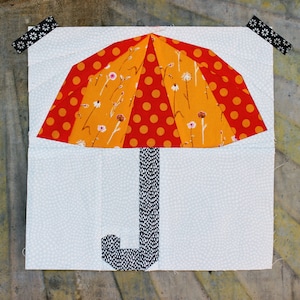 Puede incluir: Un bloque de tela con un diseño de paraguas colorido. La parte superior del paraguas tiene secciones naranjas y rojas con lunares y estampados florales. El mango es blanco y negro, sobre un fondo azul claro con puntos blancos.