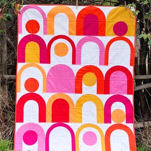 May include: A colorful quilt with a geometric pattern of arches and circles in shades of pink, orange, and red. The quilt is made of fabric and is hanging on a clothesline.