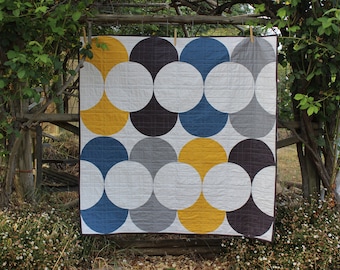 May include: A quilt with a geometric pattern of overlapping circles in shades of blue, grey, white, and yellow. The quilt is hanging on a wooden fence.